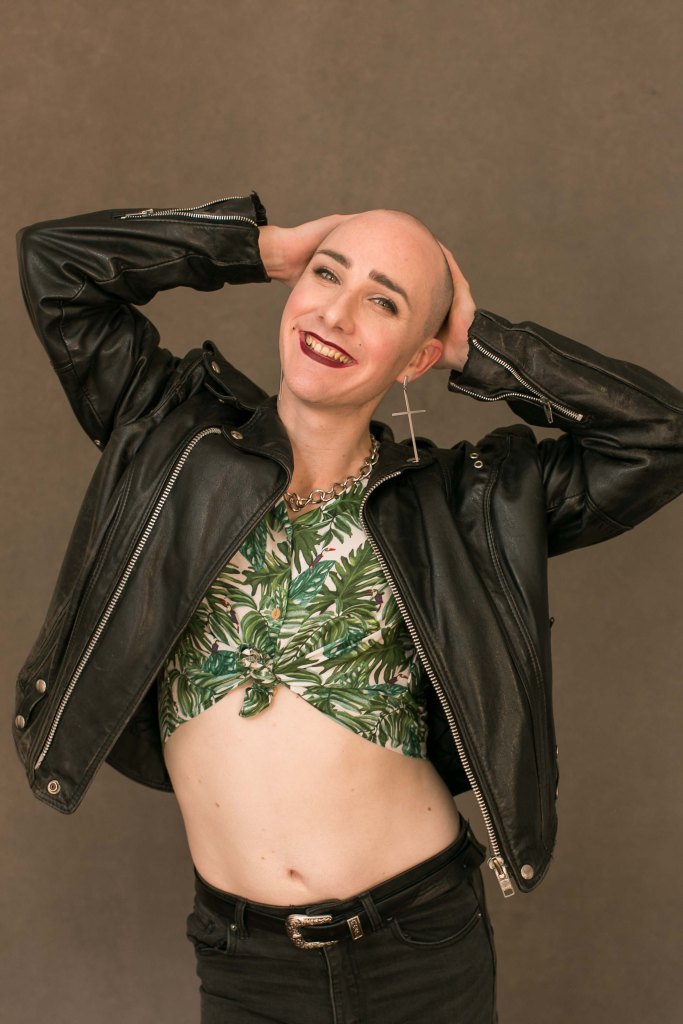 A smiling woman with a shaved head and red lipstick standing with her hands on her head in a leather jacket and fern-patterned blouse.
