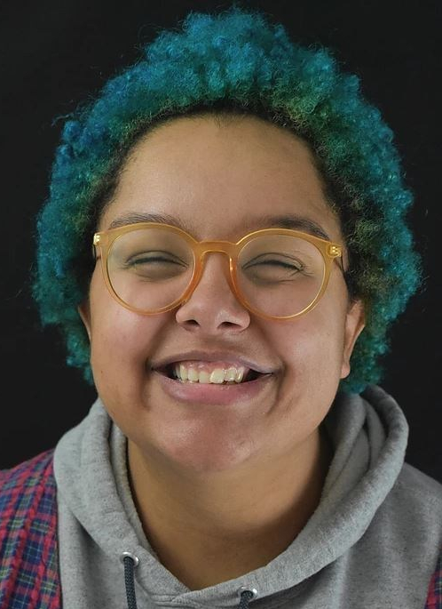 Smiling person with yellow glasses, teal and blue short curly hair and a gray hoodie.